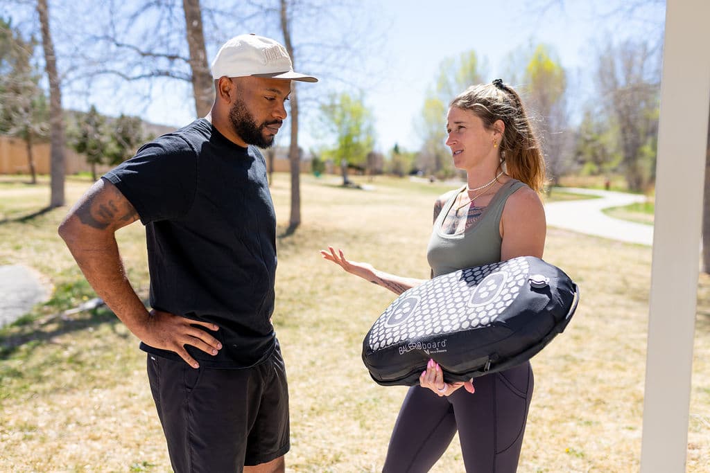 A coach holding a BALEROboard under one arm, talking with a practitioner outdoors