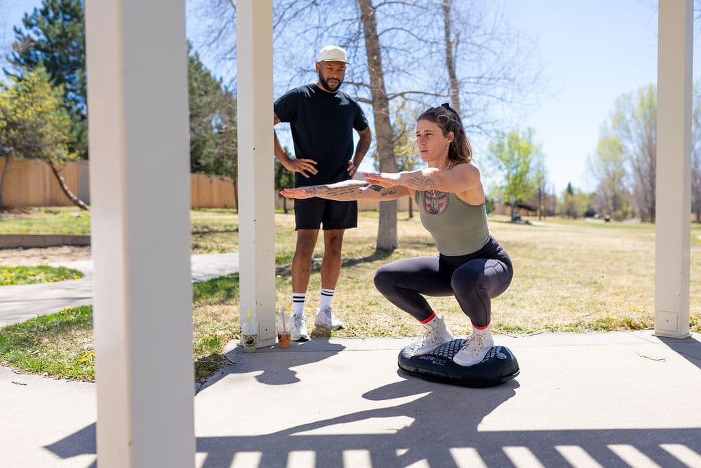 A coach watching while a client holds a squat on a BALEROboard under a pergola