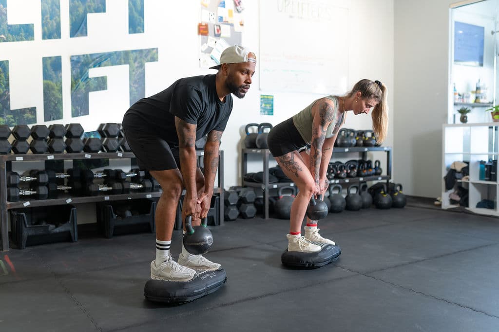 Two athletes mid-hinge with kettlebells, each standing on a BALEROboard in a gym