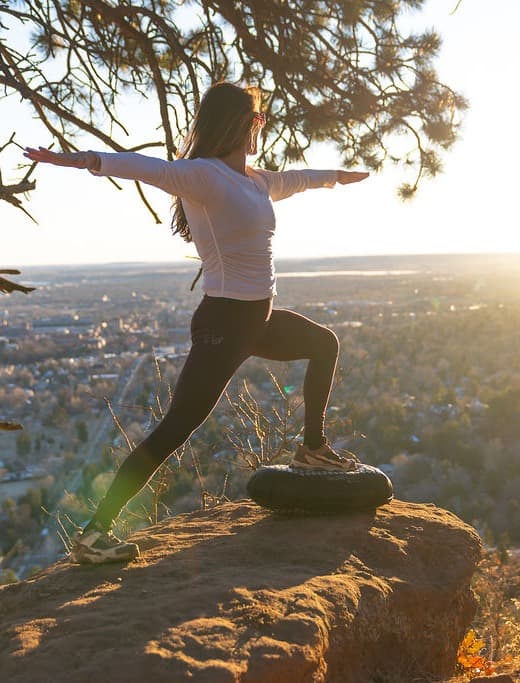 A woman holding warrior pose on a BALEROboard, on a rock above Boulder at golden hour