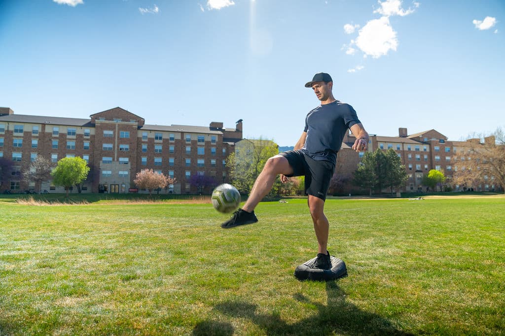 A man balancing on one leg on a BALEROboard in a field, juggling a soccer ball with the other foot